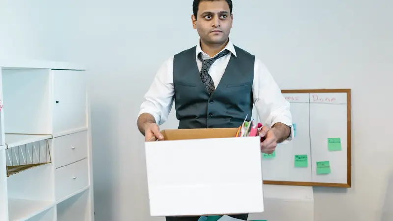 Homme en gilet et chemise tenant un carton rempli de dossiers dans un bureau, devant un tableau blanc
