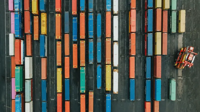 Vue aérienne de conteneurs maritimes multicolores alignés sur un parc de stockage, avec un engin de manutention rouge