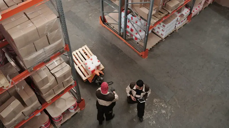 Vue plongeante sur deux magasiniers et un transpalette entre des rayonnages chargés de cartons sur palettes