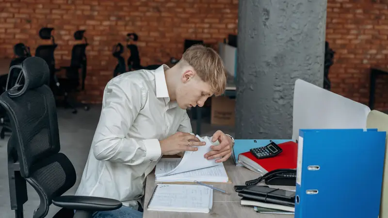 Jeune homme feuilletant une liasse de documents imprimés à un bureau, près d'un classeur bleu et d'une calculatrice