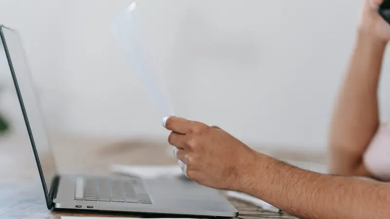 Main tenant une feuille imprimée à côté d'un ordinateur portable ouvert sur une table en bois