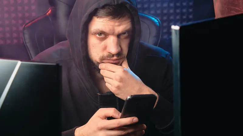 Homme en sweat à capuche tenant un smartphone, assis entre deux écrans dans une pièce sombre