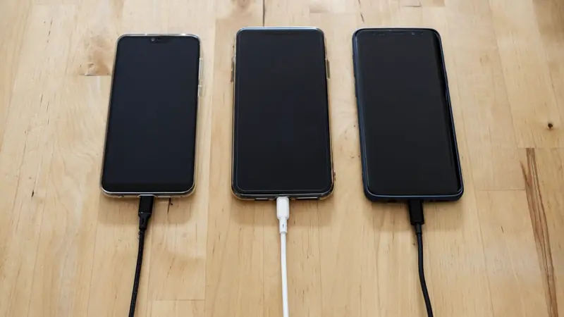 Trois smartphones posés sur une table en bois, chacun relié à un câble USB