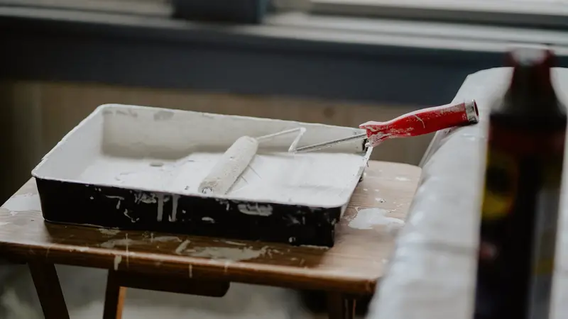 Rouleau à peinture posé dans un bac de peinture blanche sur un tabouret en bois taché